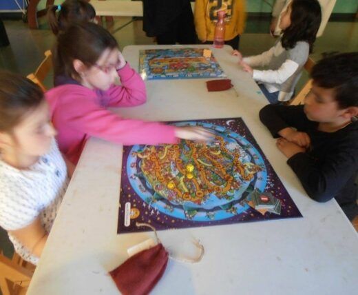 Los niños se reunieron alrededor de una mesa jugando un juego de mesa, centrándose en el colorido tablero del centro. Una chica vestida de rosa extiende su mano para mover una pieza.