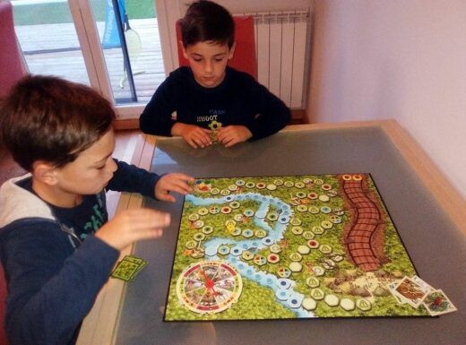 Dos niños participaban en un juego de mesa en una mesa, concentrados en planificar sus próximos movimientos. El tablero de juego presenta caminos coloridos y varias fichas de juego.